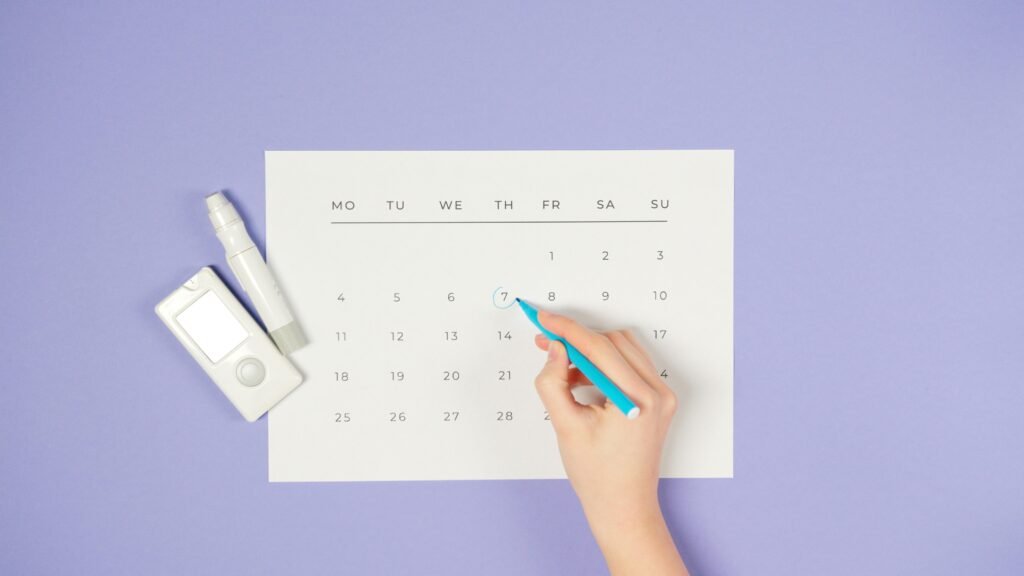pexels-photo-6823618-6823618 A hand marking a date on a calendar next to a glucometer on a purple background.