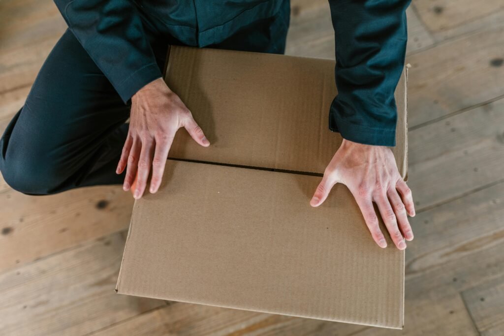 pexels-photo-7464646-7464646 Close-up of hands holding a cardboard box on a wooden floor, captured from above.